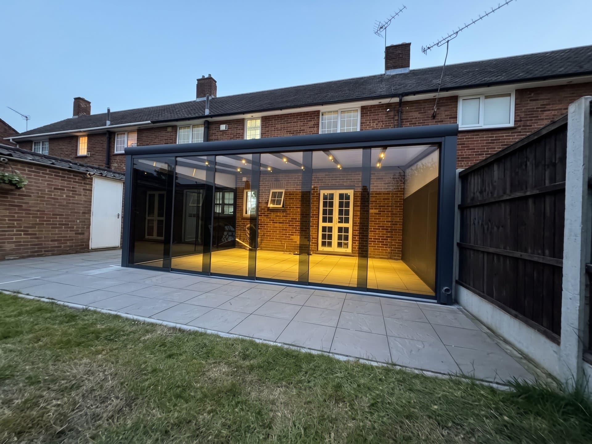 Enclosed anthracite veranda with sliding glass doors, a polycarbonate side wedge, an aluminium side wall and illuminated roof rafters.
