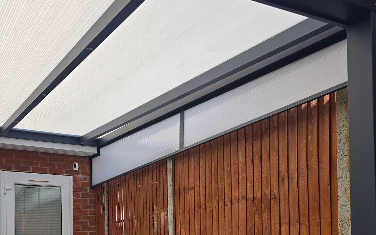 Polycarbonate side wedge beneath a sloping veranda roof, above a timber boundary fence.